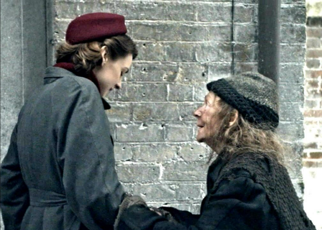 A woman in a red hat and gray coat faces an older woman in a knit hat and dark shawl in front of a brick wall. They are holding hands and smiling at each other.