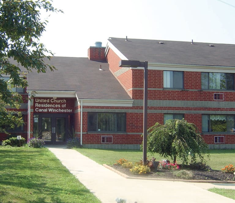 A two-story brick building with a sign that reads "United Church Residences of Canal Winchester" stands as a beacon of affordable housing in Canal Winchester, Ohio. The property features a well-maintained lawn, a paved walkway, and a landscaped garden.