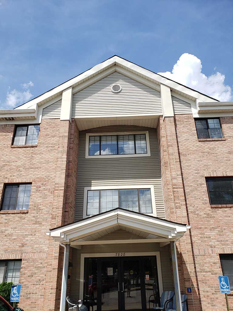 A three-story brick and siding building with a covered entrance, featuring handicap accessible signs near the front, symbolizes the charm of affordable housing in Indianapolis. The blue sky with some clouds above adds a serene backdrop to this Indiana gem.