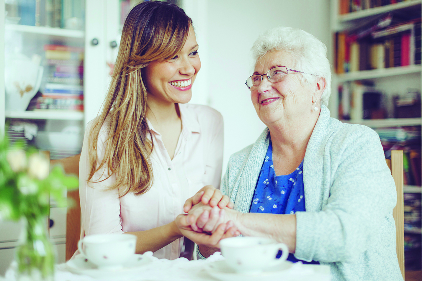 A young woman and an elderly woman sit together, smiling and holding hands, with tea cups on the table in front of them. This heartwarming scene highlights UCH's growth beyond housing to services, offering assistance wherever you live.