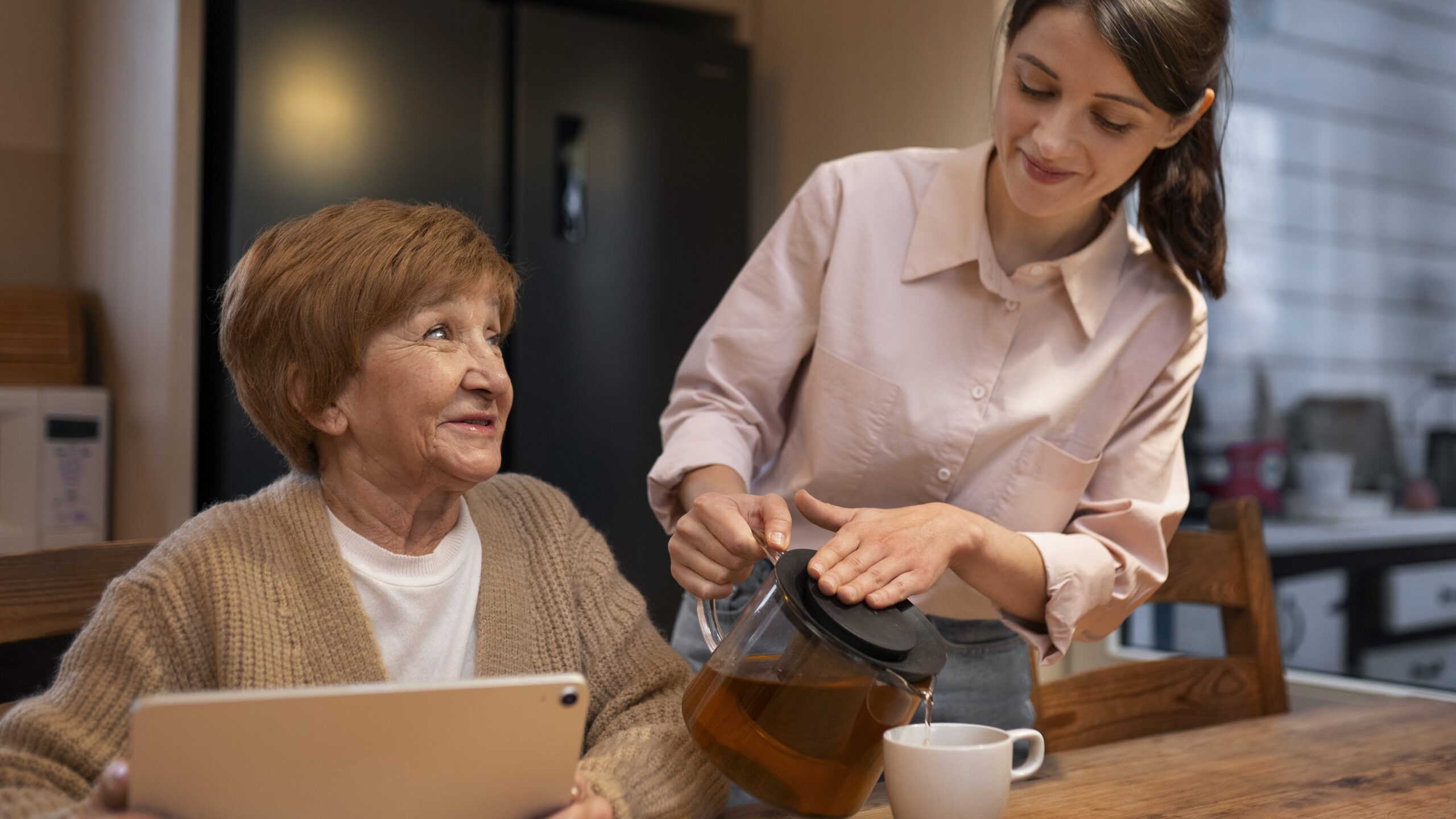 An elderly woman with a tablet and a younger woman pouring tea into a cup are seated at a wooden dining table in a kitchen setting.