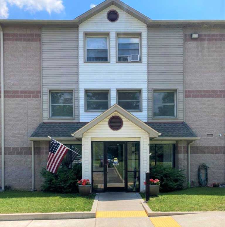 A three-story building entrance with a small overhang, displaying an American flag, and two flowerpots on either side of the doorway. The beige and brick-sided steps lead up to the affordable housing in Lakewood, New York.