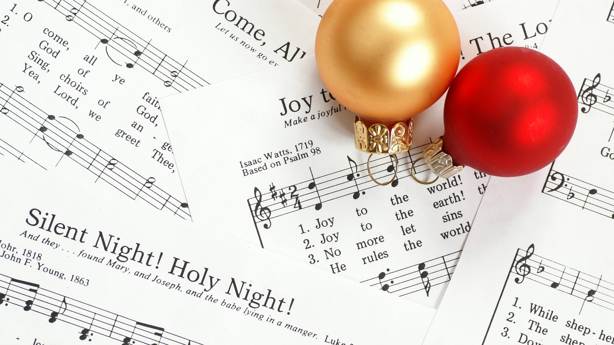 A collection of Christmas carol sheet music, including "Silent Night!" and "The First Noel," with two Christmas baubles, one red and one gold, resting on top.