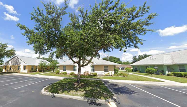 Affordable housing in Orlando, Florida, featuring a residential area with one-story houses, a central tree, and an empty parking area under a blue sky with scattered clouds.