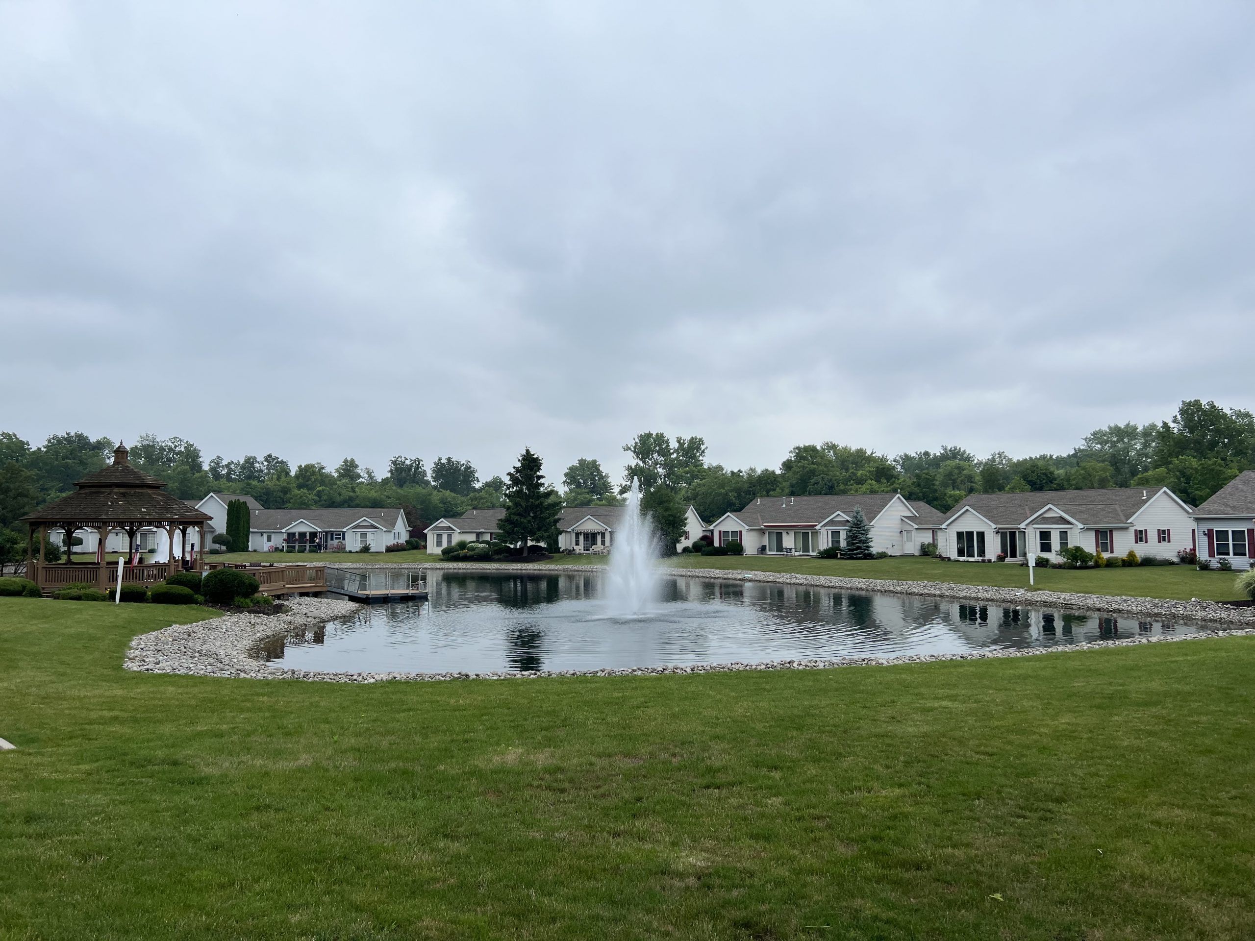 A residential community, now rebranded as The Parkvue Community on Catawba Island by United Church Homes, features houses surrounding a tranquil pond with a central fountain and a charming gazebo on the left.