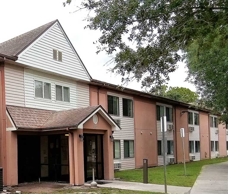 A two-story building with a sloped roof, tan and beige exterior, and several windows serves as affordable housing in Immokalee, Florida. The entrance features a small overhang, and trees are visible around the building.