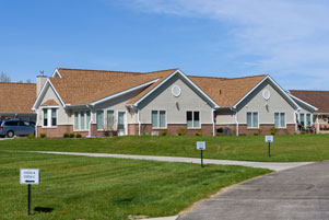 A single-story, multi-room residential building with a tan roof, gray walls, and white-trimmed windows sits on a well-maintained grassy area with signs along a paved walkway.