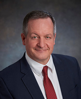 A man in a dark suit, white shirt, and red tie smiles confidently at the camera, exuding leadership against a gradient grey background.