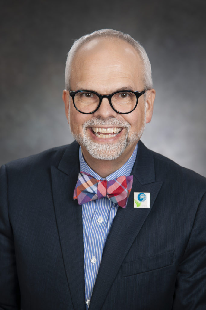 A man wearing glasses, a suit, and a colorful bow tie exudes leadership as he smiles confidently against a dark gradient background.