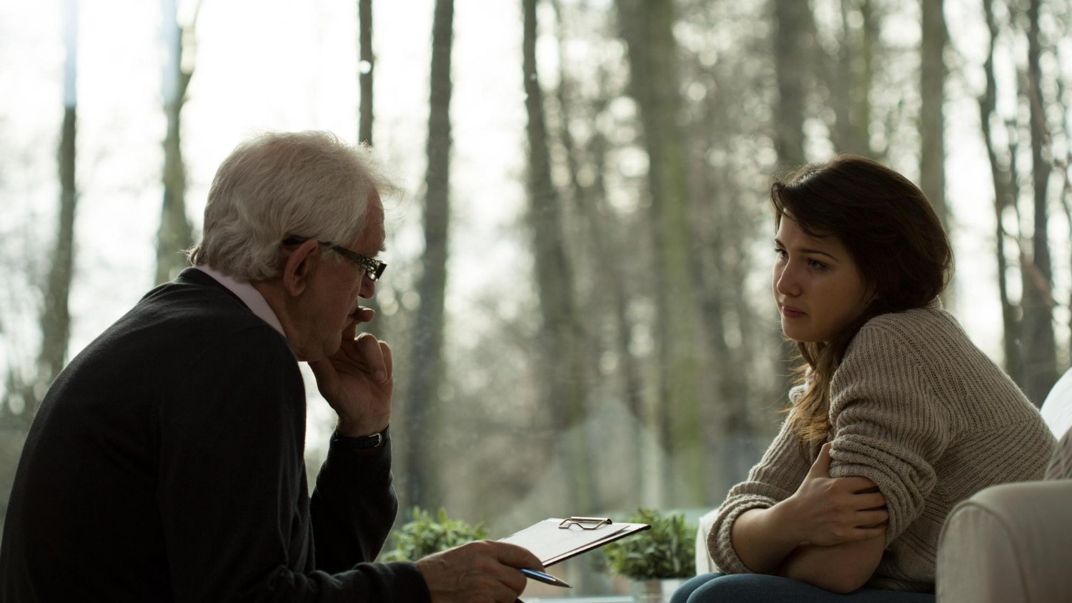 An older man with glasses and a clipboard talks to a young woman sitting with arms crossed; they are indoors by a window with a forest view.