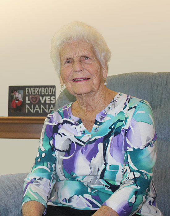 Elderly woman with white hair sitting on a blue chair, wearing a floral blouse. A sign on a shelf behind her reads "Everybody Loves Nana.