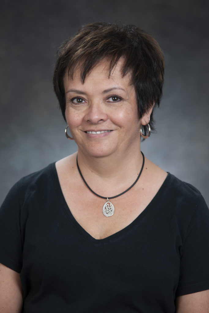 A woman with short dark hair, wearing a black top and a necklace with a pendant, smiles at the camera against a gray background, exuding leadership.