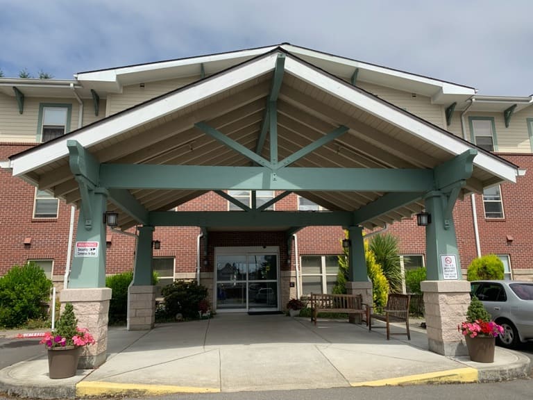 The image shows the entrance of an affordable housing building in Tacoma, Washington, featuring a brick exterior and green wooden canopy. Flower pots, benches, and a glass door adorn the entrance, while a "No Smoking" sign is visible on a pillar.