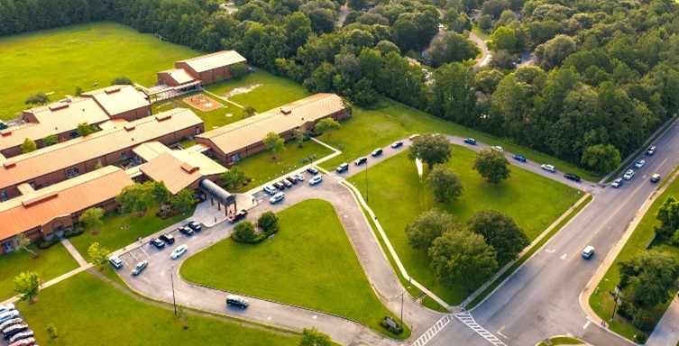 Aerial view of a school or large building with red roofs, parking lot, grassy areas, and multiple cars lined up on a driveway. Roads and trees surround the property.