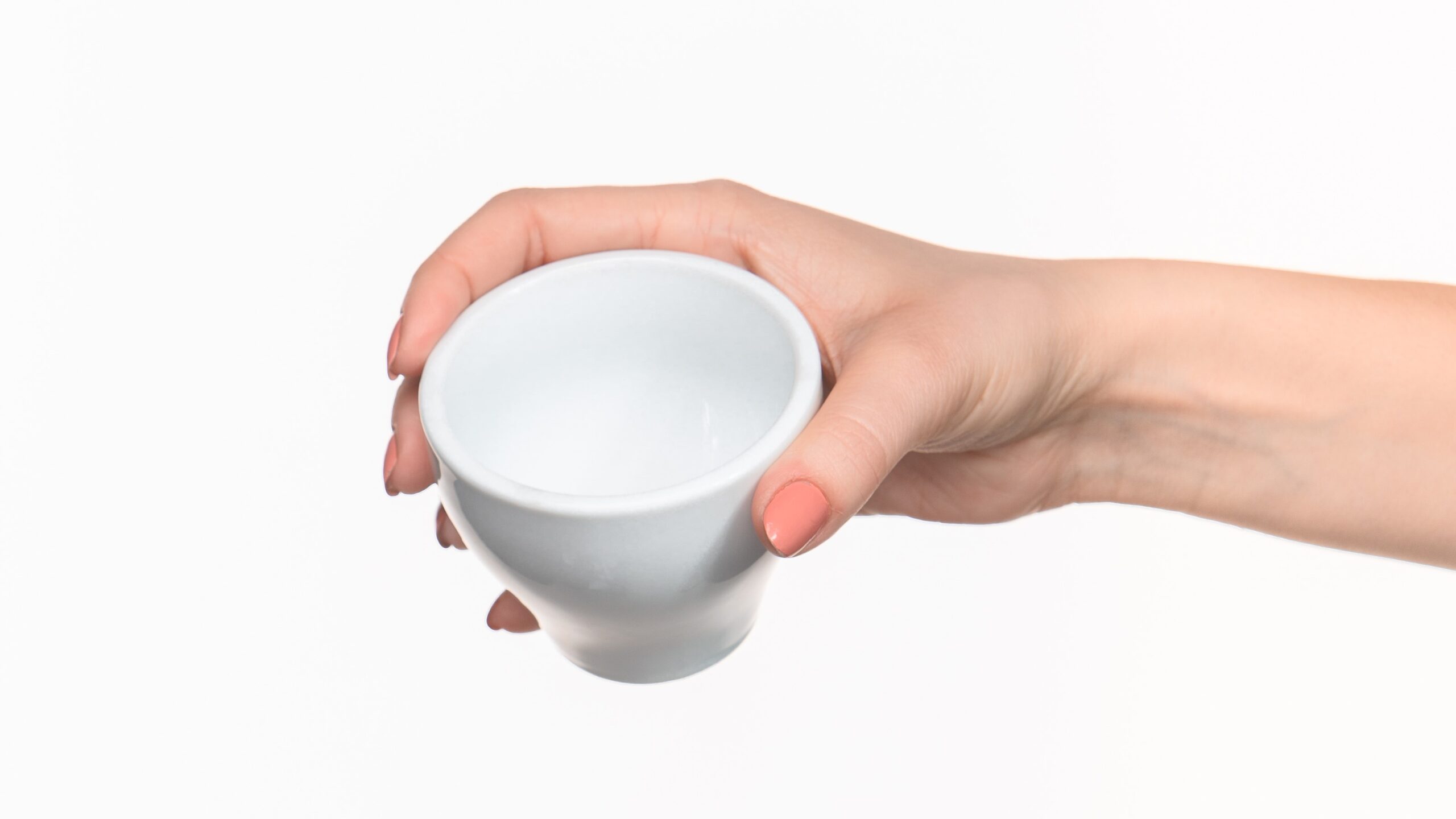 A hand with pink painted nails is holding a small white ceramic bowl against a white background.