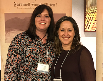 Two women smiling and standing together indoors, both wearing conference name tags. A sign in the background reads, "Farewell South Africa!.