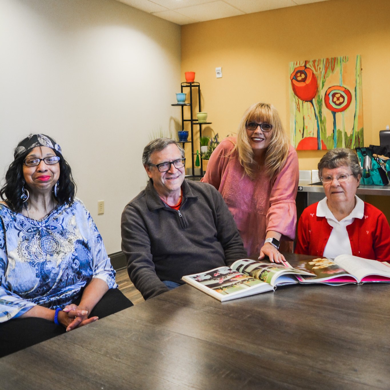 Four individuals are seated and standing around a table, looking at open books in a well-lit room. The ambiance reflects abundant living, with artwork and decorative items enriching the background.