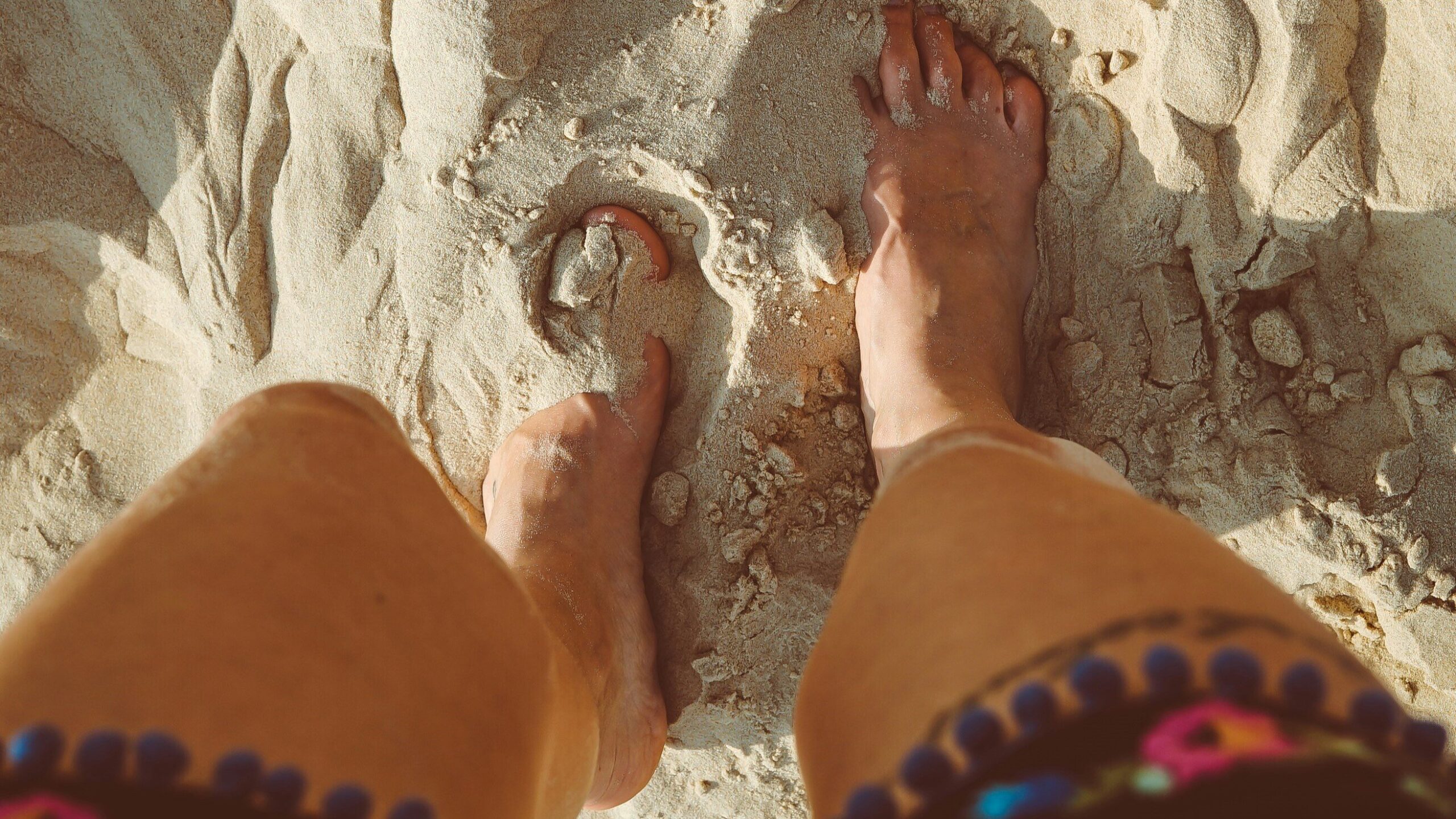 Person standing on sandy beach, feet partially buried in the sand. Detailed view of legs and feet, with the person wearing colorful, embroidered shorts.