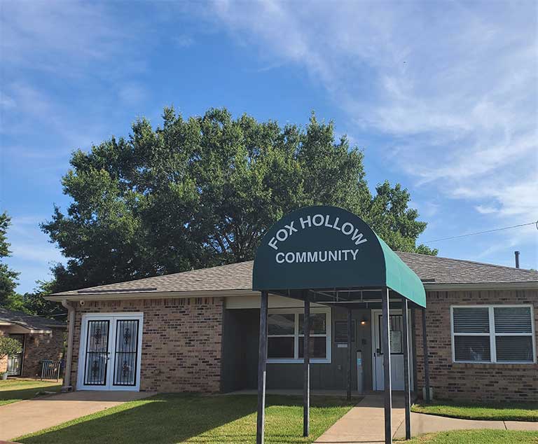 A single-story brick building with a green awning that reads “Fox Hollow Community” at the entrance. Large trees are in the background under a clear blue sky, providing a picturesque setting for this affordable housing in Covington, Tennessee.