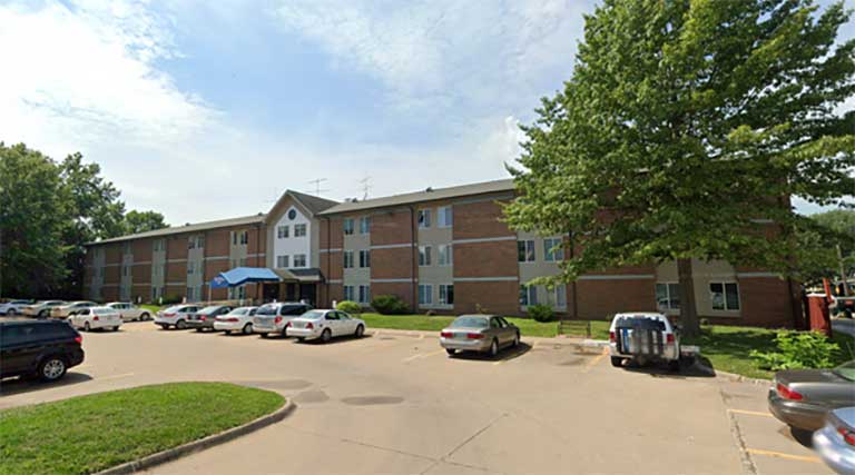 A three-story brick apartment building with a blue awning over the entrance, offering affordable housing in Burlington, Iowa. It is surrounded by trees, and a parking lot with several cars is located in front.