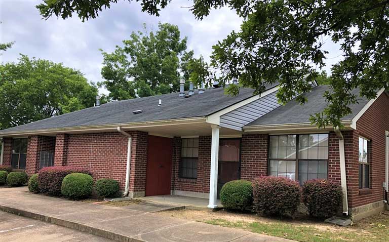 Single-story brick building with a dark shingle roof, white trim, and a front porch, surrounded by manicured bushes and trees; this charming property offers affordable housing in Columbus, Mississippi.