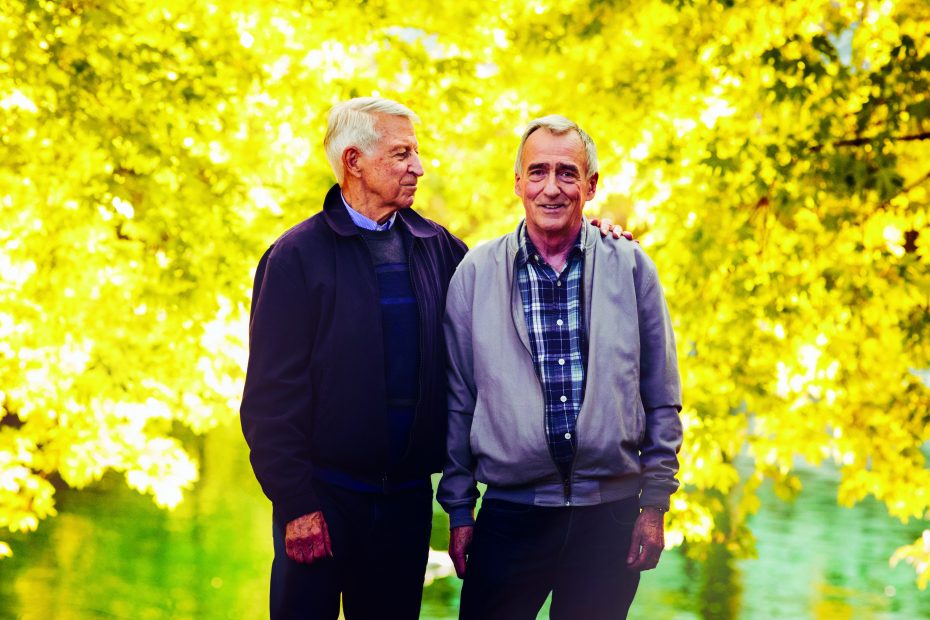 Two elderly men standing outdoors in front of a vibrant yellow-green tree. One man has his hand on the other's shoulder. Both are smiling.