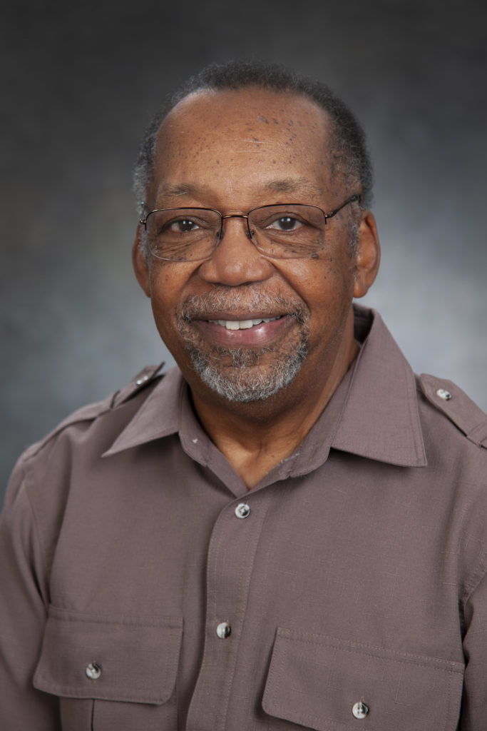 A man with glasses and a gray beard, exuding leadership in a light brown button-up shirt, smiles at the camera against a dark, neutral background.