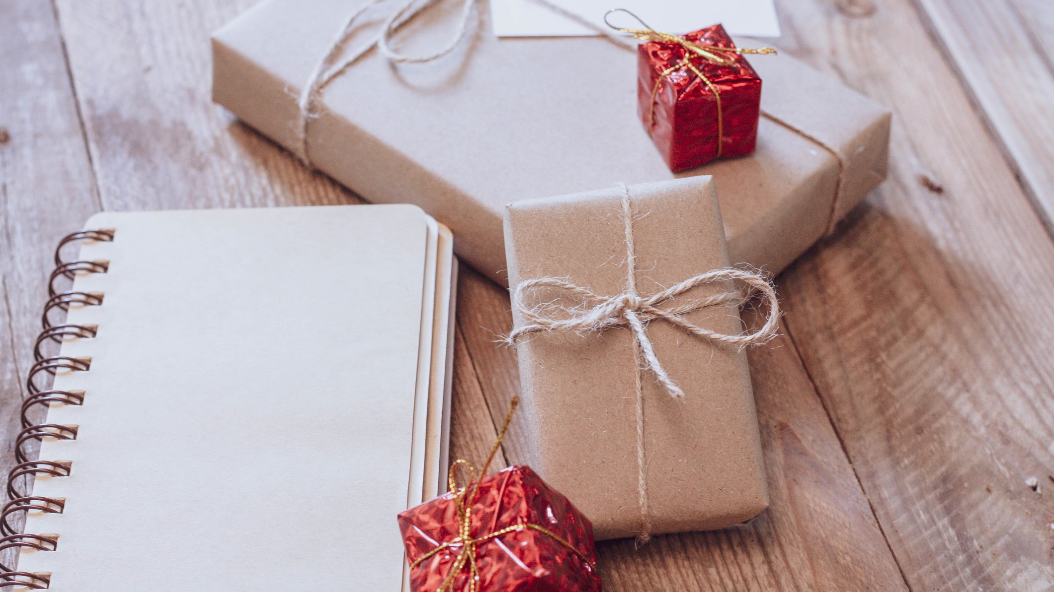 Three brown paper-wrapped gifts and a notebook are placed on a wooden surface. Two small red-wrapped presents are also shown.