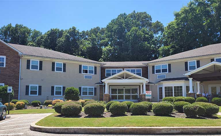 A two-story beige building with white trim, surrounded by lush green bushes and trees, under a clear blue sky. A covered entrance is at the center with a "For Sale" sign on the front lawn, offering an opportunity for affordable housing in Covington, Tennessee.