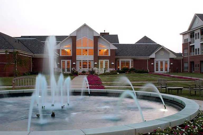 A large brick building with illuminated windows stands behind a circular fountain with multiple water jets. The fountain is surrounded by manicured lawns and pathways.