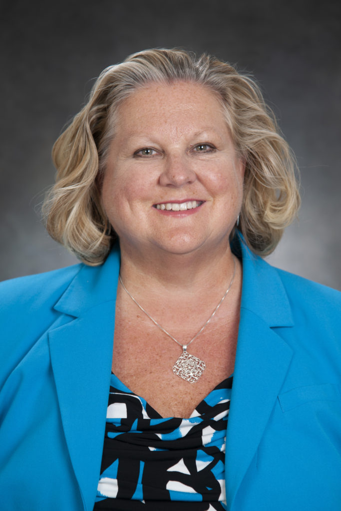 A woman with short blonde hair, wearing a blue blazer and a patterned blouse, smiles at the camera against a grey background, embodying the professional spirit of our dedicated fundraising team.