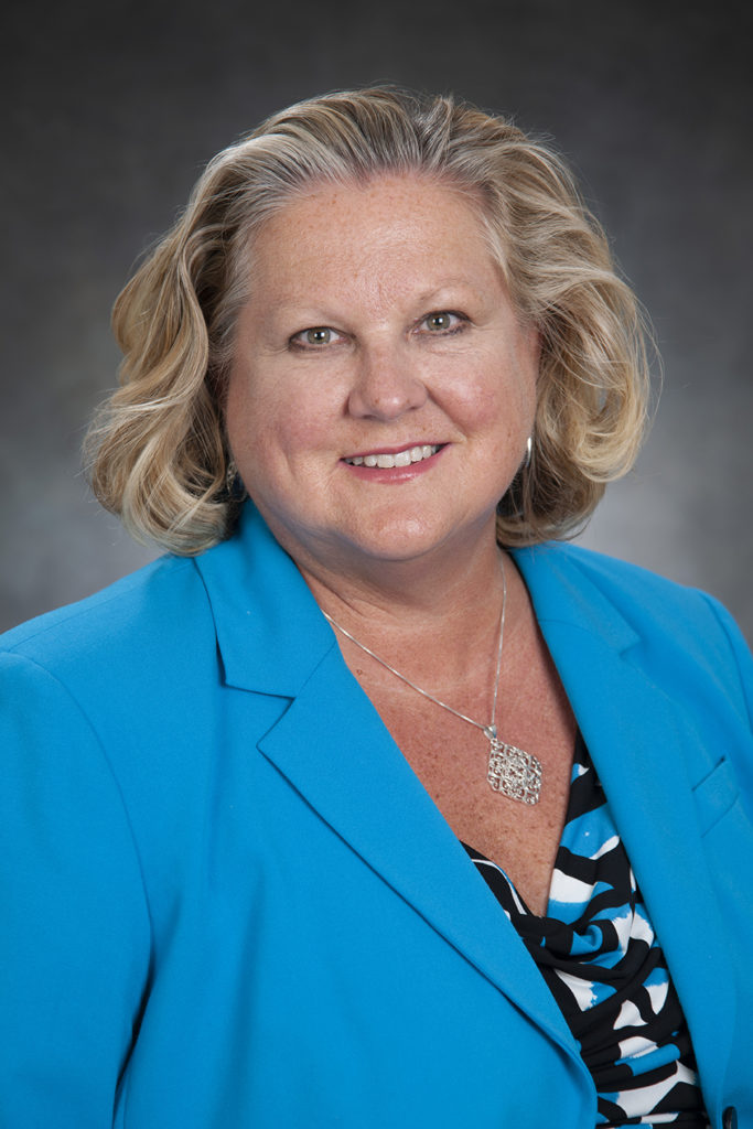 A woman with blonde hair wearing a blue blazer and a patterned blouse smiles at the camera, exuding leadership. She has a silver necklace and earrings, set against a plain studio background.