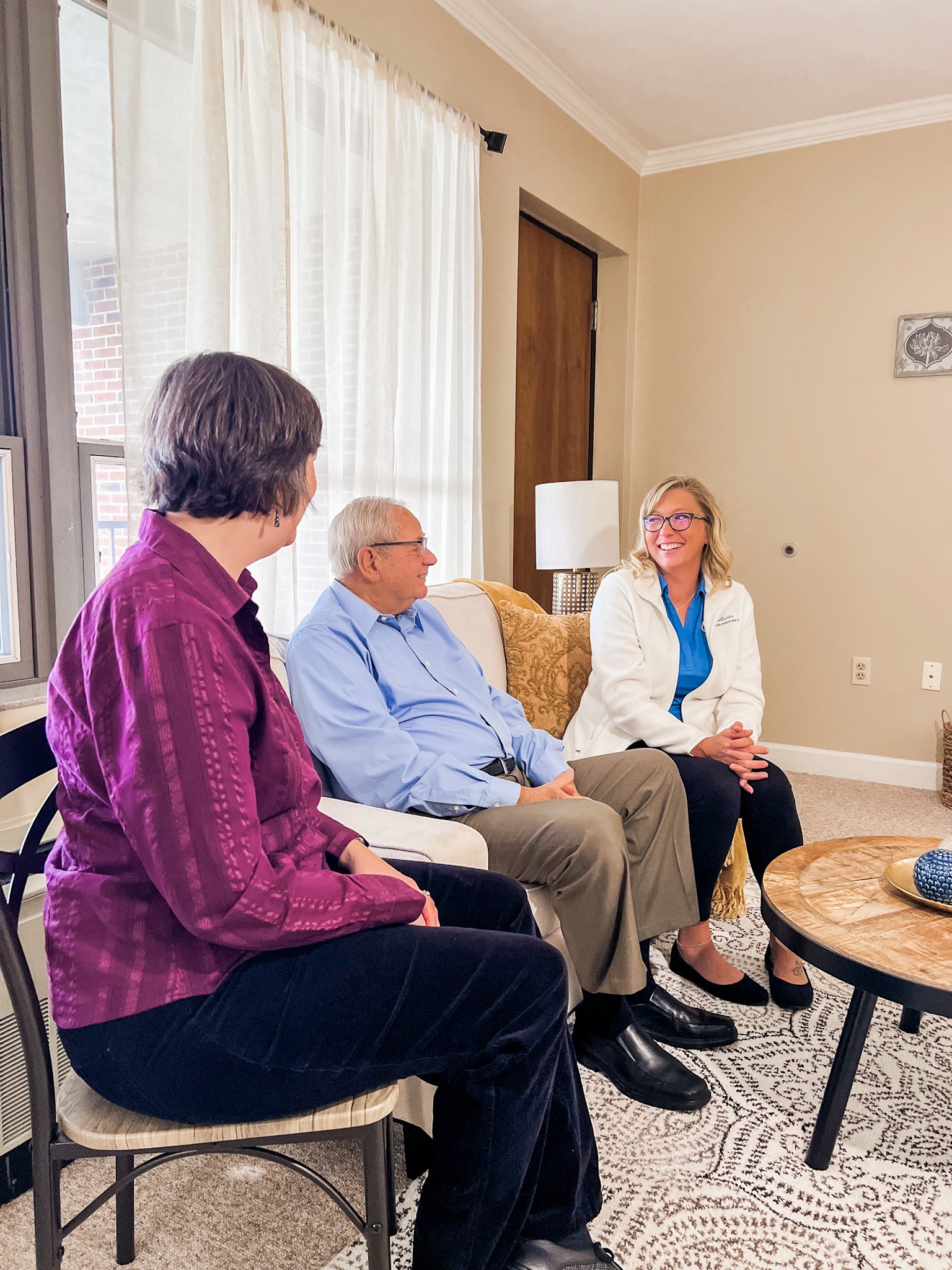 Three people sitting and conversing in a well-lit room. A man and a woman are seated on a couch, facing another woman in a white coat sitting in a chair. The abundant culture extends to staff, creating an atmosphere of comfort and open dialogue.