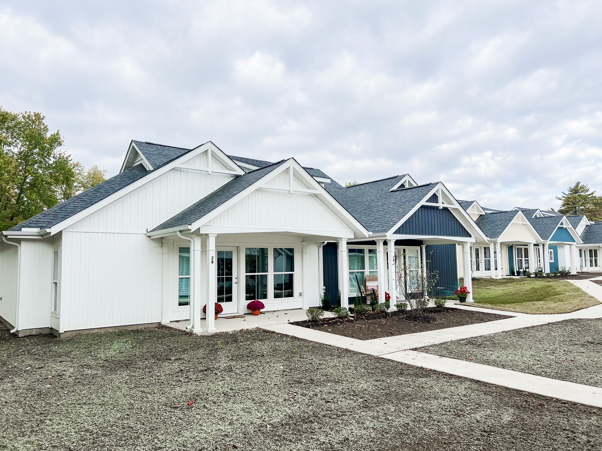 A row of modern single-story white and blue homes with well-maintained gardens and paved walkways under a cloudy sky.