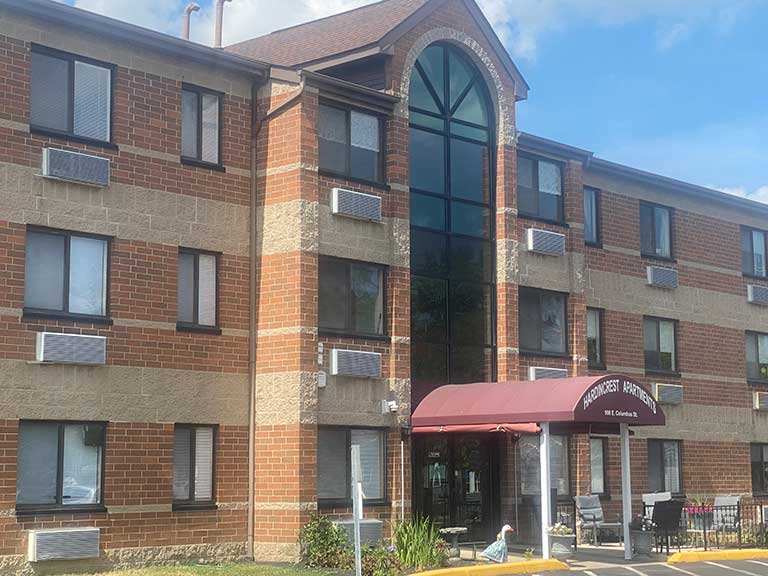 Three-story brick and stone building with a maroon awning labeled "Woodwest Apartments" in Kenton, Ohio. The building offers affordable housing and features several windows, air conditioning units, and a glass entrance with outdoor seating.