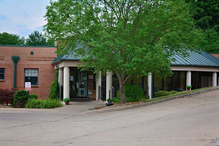 A single-story brick building with green trim and a covered entrance area, surrounded by trees and greenery, with a sloping driveway in the foreground.