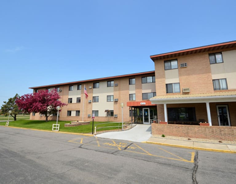 A three-story building with a beige and brown exterior in Long Prairie, Minnesota, offers affordable housing. An entrance canopy greets visitors alongside an American flag and blooming tree. The access road in front has clear markings and parking spaces under a bright blue sky.