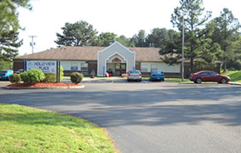 A one-story building with a sign reading "Hollyview Place" in front, surrounded by trees and parked cars. Several people are visible near the entrance of this affordable housing complex in Holly Springs, Mississippi.