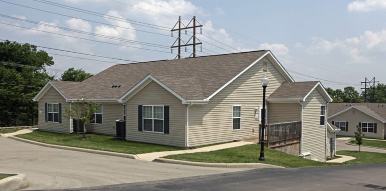 A single-story beige house with white trim, set on a sloping plot in Dayton, Ohio, electrical powerlines in the background, and a lamppost near the pathway. This property exemplifies affordable housing in Dayton, offering both charm and accessibility.