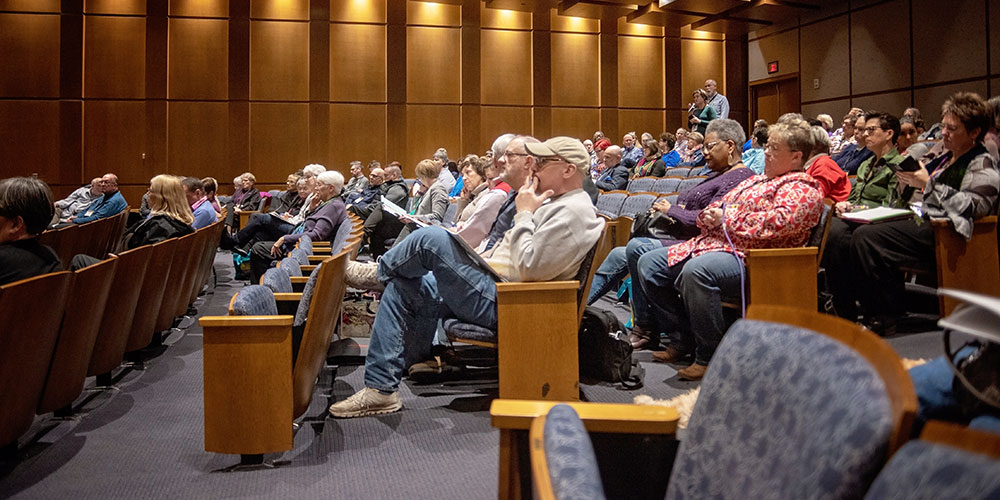 A diverse group of people seated in a wooden auditorium watching a presentation, with some taking notes.