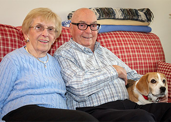 An elderly couple sits on a red checkered couch with a beagle beside them. The woman wears a blue sweater, and the man wears glasses and a checkered shirt. A folded blanket is in the background.