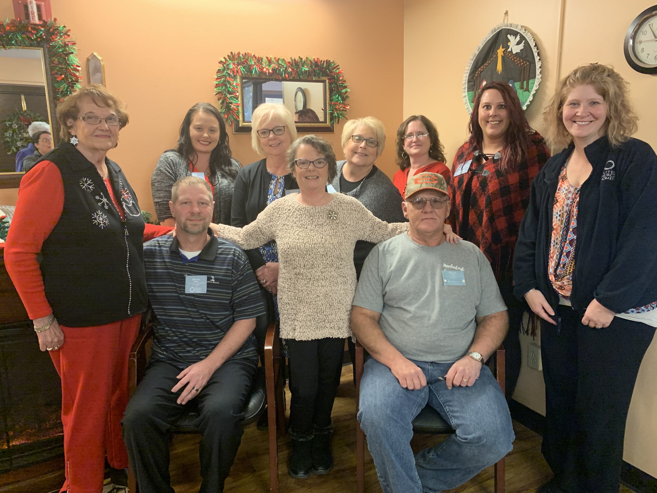 A group of ten people, some seated and some standing, pose for a photo in a decorated room with holiday garlands and a clock on the wall, celebrating Hardincrest's 30th anniversary.