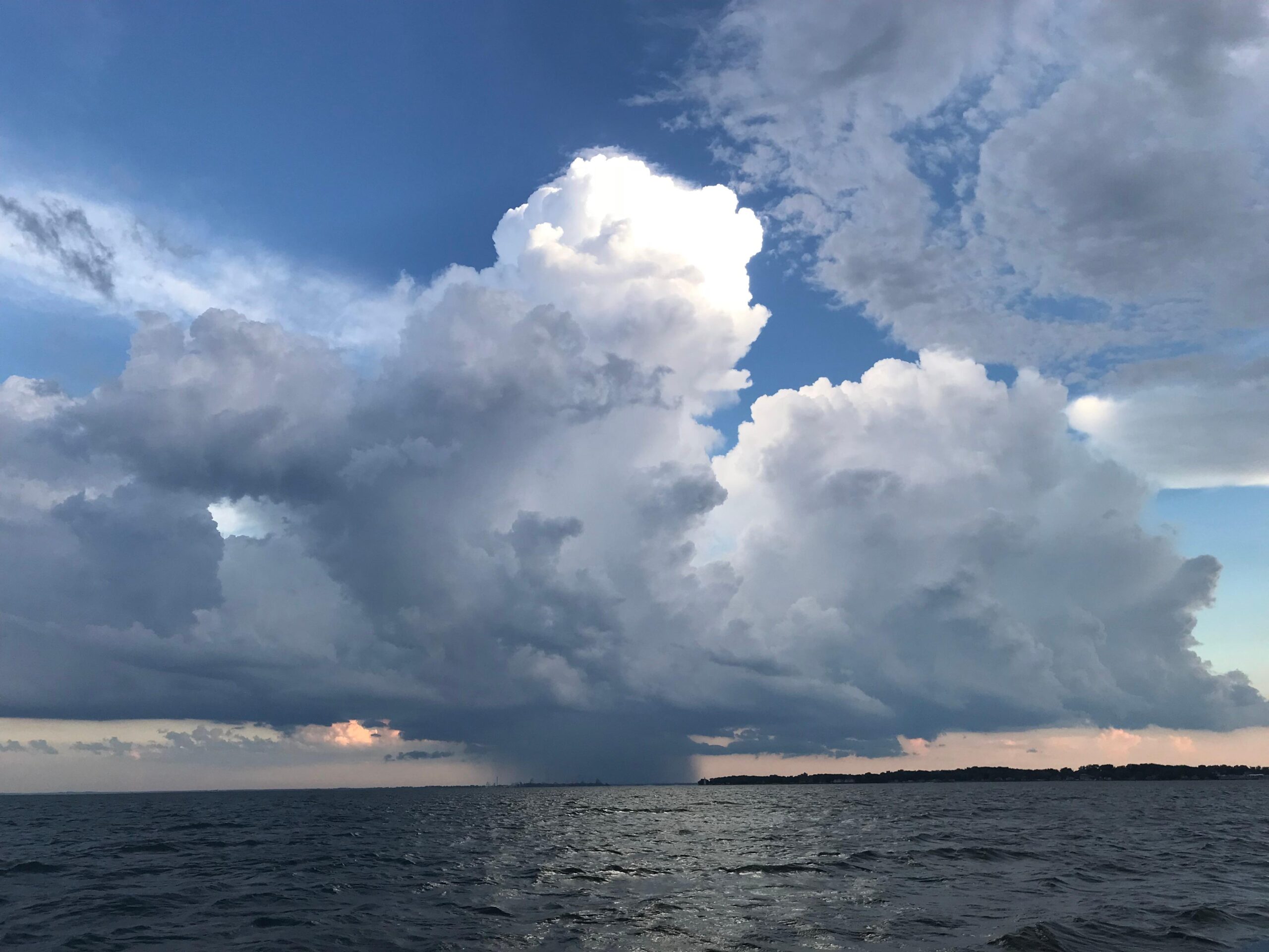 A large cumulonimbus cloud towers over the horizon above an expansive body of water under a blue sky with other smaller clouds.