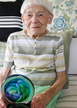 Elderly person with short white hair, wearing a striped shirt, sitting on a bed with patterned pillows, holding a colorful glass orb.