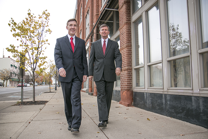 Two men in business suits walk on a city sidewalk near a brick building on a clear day, discussing plans for senior living communities.