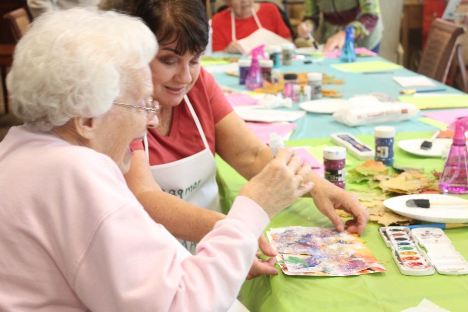 Two elderly women sitting at a table covered in art supplies, working on a watercolor painting together during a group activity.