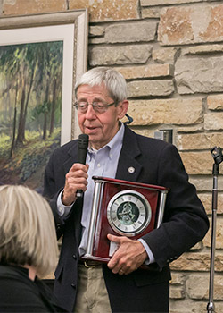 An elderly man in a dark blazer holds a clock and speaks into a microphone at an indoor event. A painting and a stone wall are in the background.