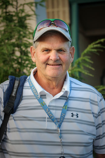 A man wearing a gray cap, sunglasses on his head, and a striped polo shirt with a lanyard around his neck stands outdoors with foliage in the background.