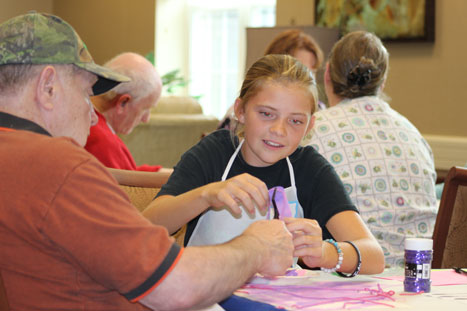 A girl and an elderly man are engaged in a craft activity at a table, surrounded by other people. She is holding a piece of yarn while he assists her. Both appear focused on the task.