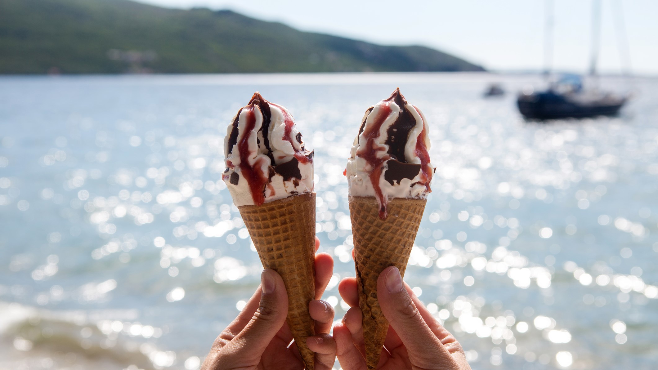 Two hands holding ice cream cones with chocolate and strawberry syrup, ocean and boats in the background.